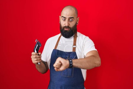 Téléchargez les photos : Jeune homme hispanique avec barbe et tatouages portant tablier de coiffeur tenant rasoir regardant l'heure de la montre inquiet, peur de se faire tard - en image libre de droit