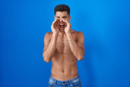 Foto de Joven hombre hispano parado sin camisa sobre fondo azul gritando furioso con las manos sobre la boca - Imagen libre de derechos