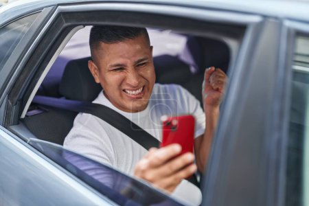Foto de Joven hispano haciendo videollamada con smartphone en el coche gritando orgulloso, celebrando la victoria y el éxito muy emocionado con el brazo levantado - Imagen libre de derechos