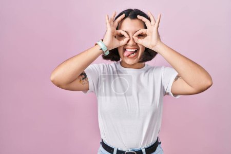 Foto de Mujer hispana joven vistiendo una camiseta blanca casual sobre fondo rosa haciendo un buen gesto como prismáticos sacando la lengua, ojos mirando a través de los dedos. expresión loca. - Imagen libre de derechos