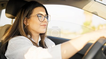 Téléchargez les photos : Jeune belle femme hispanique conduisant une voiture souriante portant des lunettes sur la route - en image libre de droit