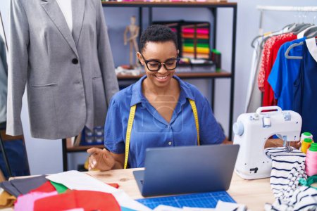 Téléchargez les photos : Femme afro-américaine tailleur souriant confiant en utilisant un ordinateur portable au magasin de tailleur - en image libre de droit