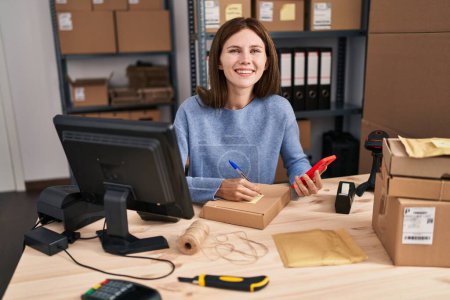 Téléchargez les photos : Jeune femme blonde ecommerce travailleur d'affaires en utilisant smartphone écriture sur le paquet au bureau - en image libre de droit