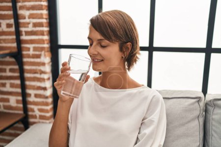 Téléchargez les photos : Jeune belle hispanique femme boire un verre d'eau assis sur le canapé à la maison - en image libre de droit