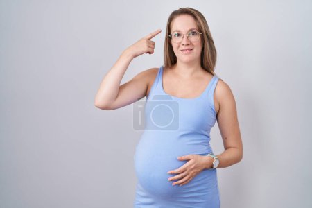 Foto de Mujer embarazada joven de pie sobre fondo blanco sonriendo señalando a la cabeza con un dedo, gran idea o pensamiento, buena memoria - Imagen libre de derechos