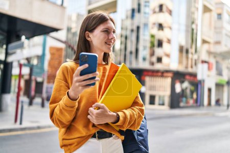 Téléchargez les photos : Jeune étudiante blonde utilisant un smartphone tenant des livres dans la rue - en image libre de droit