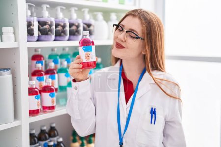 Téléchargez les photos : Jeune rousse femme pharmacien souriant confiant tenant bouteille de médicaments à la pharmacie - en image libre de droit