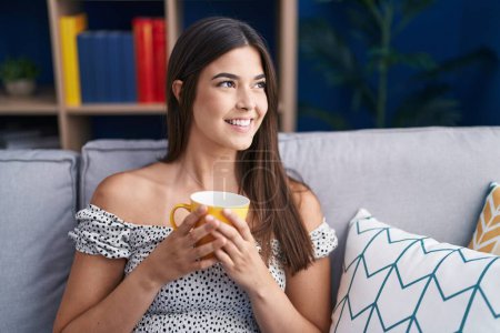 Photo for Young beautiful hispanic woman drinking coffee sitting on sofa at home - Royalty Free Image