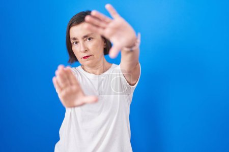 Foto de Mujer hispana de mediana edad de pie sobre fondo azul haciendo marco usando las manos palmas y los dedos, perspectiva de la cámara - Imagen libre de derechos