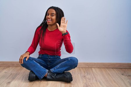 Foto de Joven afroamericano con trenzas sentado en el suelo en casa renunciando a decir hola feliz y sonriente, gesto de bienvenida amistoso - Imagen libre de derechos