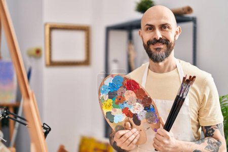 Téléchargez les photos : Jeune homme chauve artiste souriant confiant tenant pinceaux et palette au studio d'art - en image libre de droit