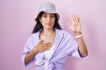 Foto de Mujer hispana joven de pie sobre fondo rosa con sombrero jurando con la mano en el pecho y la palma abierta, haciendo un juramento de lealtad promesa - Imagen libre de derechos