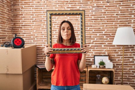 Foto de Mujer hispana joven en casa nueva sosteniendo marco vacío haciendo cara de pez con la boca y ojos entrecerrados, loco y cómico. - Imagen libre de derechos