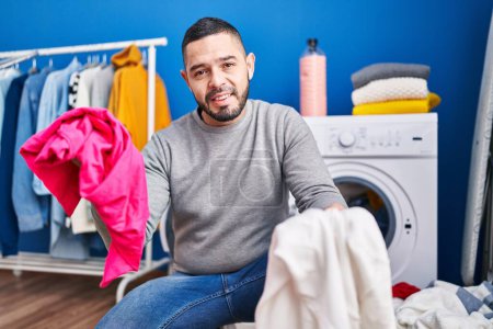 Foto de Joven latino sonriendo confiado lavando ropa en la lavandería - Imagen libre de derechos