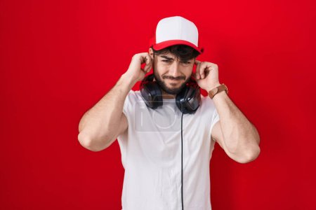 Foto de Hombre hispano con barba usando sombrero de gamer y auriculares cubriendo oídos con dedos con expresión molesta por el ruido de la música alta. concepto de sordo. - Imagen libre de derechos