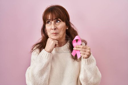 Foto de Middle age hispanic woman holding pink cancer ribbon serious face thinking about question with hand on chin, thoughtful about confusing idea - Imagen libre de derechos