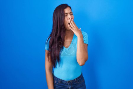 Foto de Mujer hispana joven de pie sobre fondo azul aburrida bostezando cansada cubriendo la boca con la mano. inquietud y somnolencia. - Imagen libre de derechos