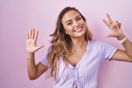 Foto de Mujer hispana joven de pie sobre fondo rosa mostrando y señalando hacia arriba con los dedos número ocho mientras sonríe confiado y feliz. - Imagen libre de derechos