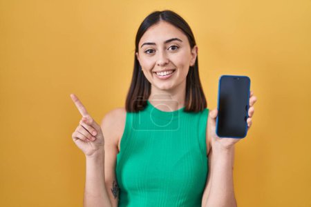 Téléchargez les photos : Fille hispanique tenant smartphone montrant écran souriant heureux pointant avec la main et le doigt sur le côté - en image libre de droit