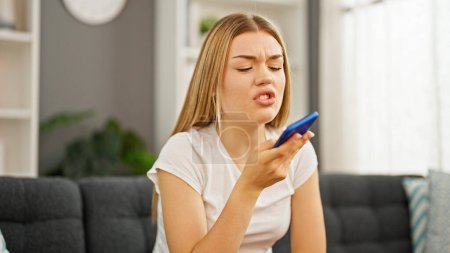 Foto de Mujer rubia joven enviando mensaje de voz por teléfono inteligente enojado en casa - Imagen libre de derechos
