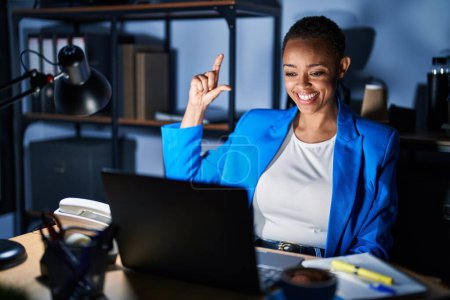 Téléchargez les photos : Belle femme afro-américaine qui travaille au bureau la nuit en souriant et en faisant des gestes confiants avec la main faisant signe de petite taille avec les doigts à la recherche et la caméra. concept de mesure. - en image libre de droit