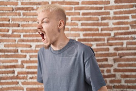 Téléchargez les photos : Jeune homme caucasien debout au-dessus du mur de briques en colère et fou hurlant frustré et furieux, criant de colère. rage et concept agressif. - en image libre de droit