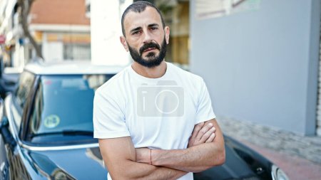 Téléchargez les photos : Jeune homme hispanique assis sur une voiture avec les bras croisés geste et visage sérieux à la rue - en image libre de droit