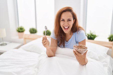 Téléchargez les photos : Brunette femme manger le petit déjeuner dans le lit souriant et riant fort à haute voix parce que drôle blague folle. - en image libre de droit