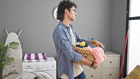Foto de Joven hombre hispano sosteniendo canasta con ropa con cara seria en la lavandería - Imagen libre de derechos
