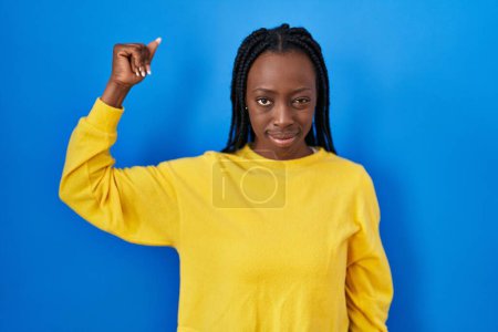 Téléchargez les photos : Belle femme noire debout sur fond bleu personne forte montrant muscle du bras, confiant et fier de la puissance - en image libre de droit