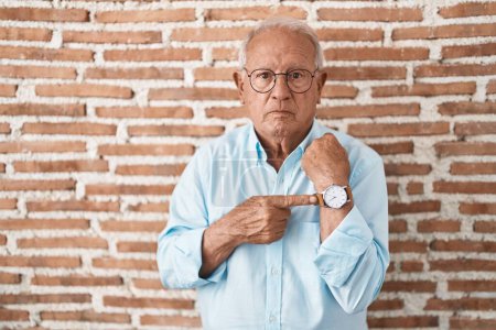 Téléchargez les photos : Homme âgé avec les cheveux gris debout sur le mur de briques à la hâte pointant pour regarder le temps, l'impatience, regardant la caméra avec une expression détendue - en image libre de droit