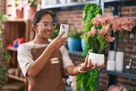 Téléchargez les photos : Jeune belle hispanique femme fleuriste faire une photo à planter par smartphone chez le fleuriste - en image libre de droit