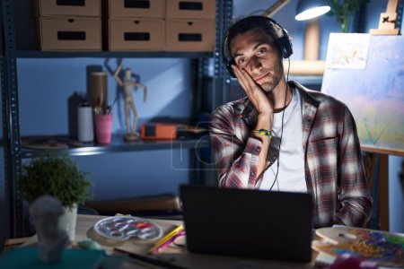 Téléchargez les photos : Jeune homme hispanique assis au studio d'art avec un ordinateur portable tard dans la nuit pensant fatigué et ennuyé par les problèmes de dépression avec les bras croisés. - en image libre de droit