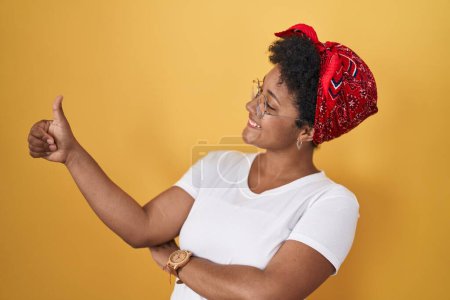 Foto de Joven mujer afroamericana de pie sobre fondo amarillo mirando orgulloso, sonriendo haciendo pulgares hacia arriba gesto a un lado - Imagen libre de derechos