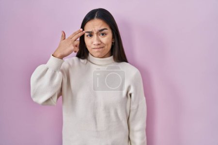 Foto de Joven mujer sudasiática de pie sobre fondo rosa disparando y matándose señalando la mano y los dedos a la cabeza como arma, gesto suicida. - Imagen libre de derechos