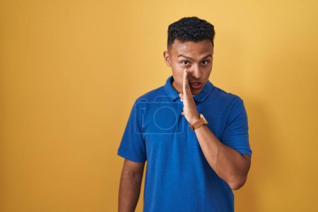 Téléchargez les photos : Jeune homme hispanique debout sur fond jaune main sur bouche racontant rumeur secrète, chuchotant conversation malicieuse - en image libre de droit