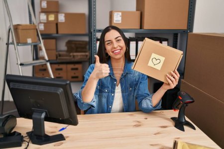Photo for Hispanic woman working at small business ecommerce holding box smiling happy and positive, thumb up doing excellent and approval sign - Royalty Free Image