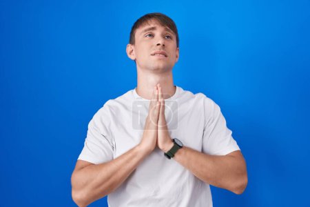 Téléchargez les photos : Homme blond caucasien debout sur fond bleu mendier et prier avec les mains ainsi que l'expression d'espoir sur le visage très émotionnel et inquiet. mendier. - en image libre de droit