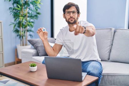 Photo for Handsome latin man holding virtual currency bitcoin using laptop pointing with finger to the camera and to you, confident gesture looking serious - Royalty Free Image
