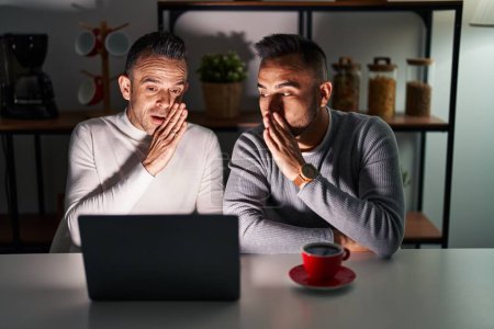 Téléchargez les photos : Couple homosexuel utilisant ordinateur portable main sur la bouche dire rumeur secrète, chuchotant conversation malicieuse - en image libre de droit