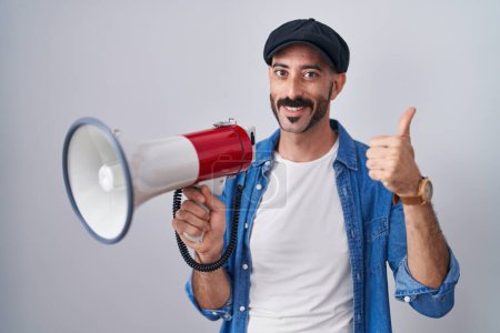 Téléchargez les photos : Homme hispanique avec barbe criant à travers mégaphone souriant heureux et positif, pouce levé faire excellent et signe d'approbation - en image libre de droit