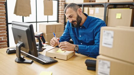 Photo for Young hispanic man ecommerce business worker talking on smartphone writing on package at office - Royalty Free Image
