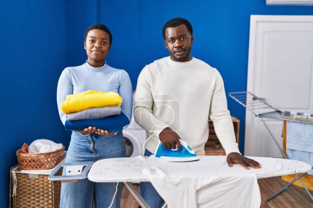 Foto de Joven pareja afroamericana planchando ropa en la lavandería relajado con expresión seria en la cara. simple y natural mirando a la cámara. - Imagen libre de derechos