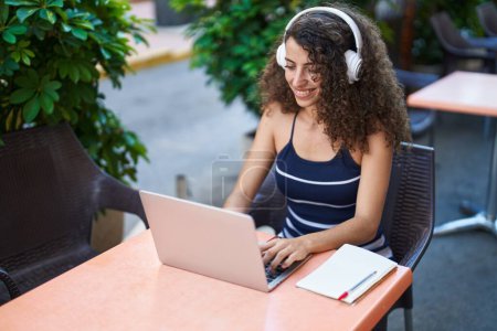 Téléchargez les photos : Jeune belle femme hispanique utilisant ordinateur portable et écouteurs assis sur la table à la terrasse du café - en image libre de droit