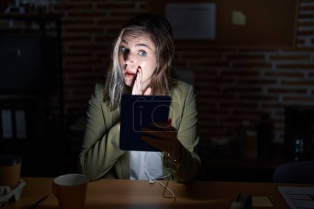 Téléchargez les photos : Blonde femme caucasienne travaillant au bureau la nuit main sur la bouche disant rumeur secrète, chuchotant conversation malicieuse - en image libre de droit
