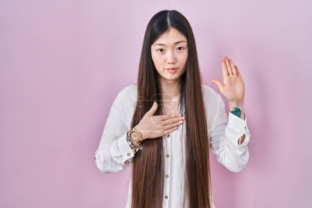 Photo for Chinese young woman standing over pink background swearing with hand on chest and open palm, making a loyalty promise oath - Royalty Free Image
