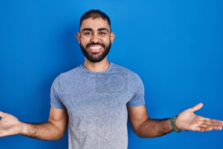 Photo for Middle east man with beard standing over blue background smiling cheerful with open arms as friendly welcome, positive and confident greetings - Royalty Free Image
