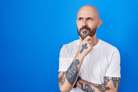 Foto de Hombre hispano con tatuajes de pie sobre fondo azul mirando confiado a la cámara sonriendo con los brazos cruzados y la mano levantada en la barbilla. pensar positivamente. - Imagen libre de derechos