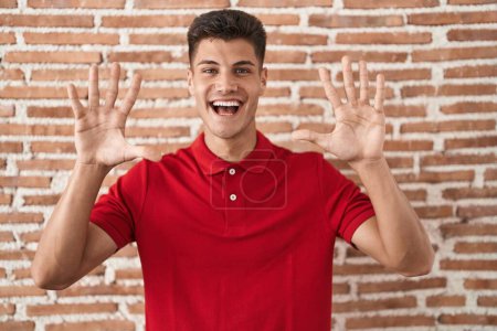 Téléchargez les photos : Jeune homme hispanique debout sur le mur de briques montrant et pointant vers le haut avec les doigts numéro dix tout en souriant confiant et heureux. - en image libre de droit
