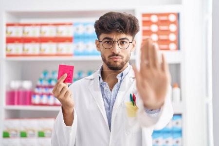 Photo for Arab man with beard working at pharmacy drugstore holding condom puffing cheeks with funny face. mouth inflated with air, catching air. - Royalty Free Image